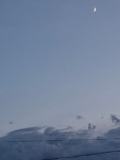 In the clear sky above Novostroyka, Russia, a crescent moon is visible in the upper right quadrant of the frame. The sky is a gradient of pale blue, transitioning to a lighter shade towards the horizon. In the lower portion of the image, dark, wispy clouds form a band across the sky. Two thin, dark power lines are strung horizontally across the lower third of the image, obscuring a portion of the clouds. The overall scene depicts an evening sky with celestial and atmospheric elements.