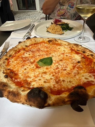 An overhead shot captures a close-up view of a pizza on a white tablecloth. The pizza is circular and topped with tomato sauce and melted mozzarella cheese, with a single green basil leaf placed in the center. The crust is slightly charred in places. To the right of the pizza is a white plate with a serving of food, including what appears to be a piece of fish coated in nuts, served over a bed of spinach, and garnished with red threads. A wine glass filled with white wine sits beside the plate. In the background, a person wearing a floral-patterned top can be seen. The setting appears to be an outdoor dining area, with cobblestone pavement visible behind the table. The scene suggests a meal at a restaurant in San Giovanni, Italy, given the distinct architectural elements suggestive of Italian regions and the presence of food and drink commonly found in Italian cuisine.