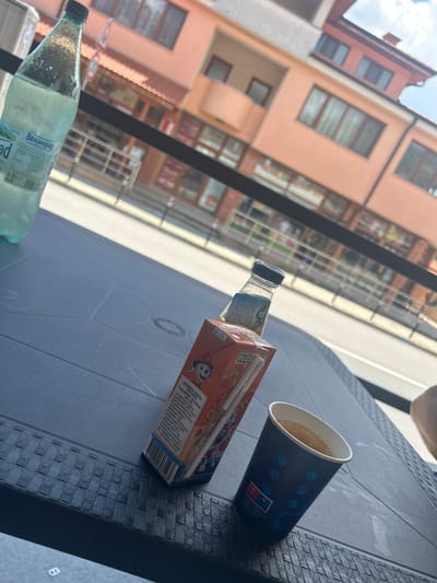 On a dark grey, textured plastic table in Rozino, Bulgaria, several drinks are placed in view of a building with peach-colored walls. To the left, a large, clear plastic bottle of "Велинград" mineral water stands upright, its contents partially visible. Next to it, closer to the center, is a small, clear bottle with a dark cap. In front of this is a small, orange juice box with a straw. To the right of the juice box is a dark blue paper cup with a white rim, filled with a light brown beverage, possibly coffee. The background shows a street scene with a building featuring multiple windows and balconies. The sky above is bright and appears cloudy.