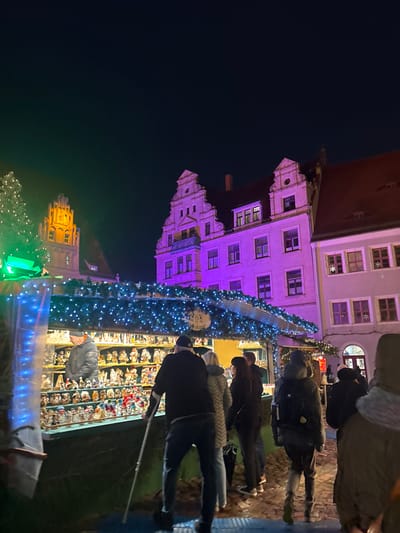 A nighttime scene depicts a European Christmas market. A tall, illuminated tree stands next to vendor stalls displaying figurines. People browse the merchandise. Ornate buildings with intricate architectural details are illuminated in purple and gold lighting, creating a festive and vibrant atmosphere. The dark sky contrasts with the brightly lit market.