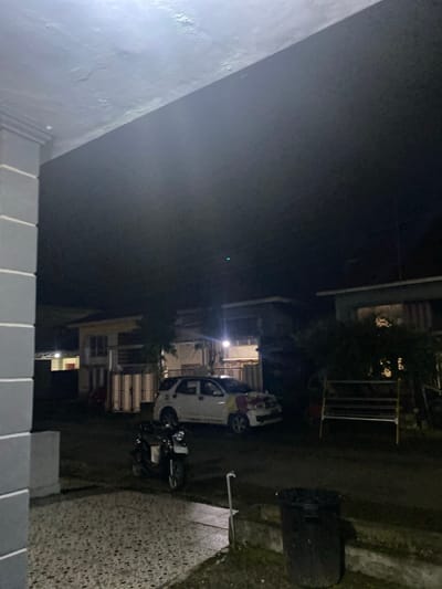 The image depicts a nighttime scene in Walennae, Indonesia. The foreground is dominated by a light-colored, speckled paved area and a portion of a building's gray, textured wall on the left. Beyond this, a dark asphalt road is visible. On the road, a dark-colored scooter is parked, and further down, a white SUV is positioned. The SUV appears to be decorated with red and yellow elements on its front.

Several residential buildings are present in the background. One building, with a light-colored facade and dark windows, is situated centrally. To its left, another structure with a reddish door and white vertical panels is partially visible. To the right of the SUV, a yellow and metal frame structure, possibly a drying rack or play equipment, stands out against the darkness. The environment appears to be a residential area, with some vegetation visible near the buildings. The overall lighting suggests artificial illumination from nearby structures. The sky is dark, indicating it is nighttime.