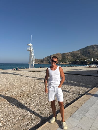 A man stands on a pebble beach, wearing a white tank top and white shorts. He is also wearing sunglasses and light-colored clogs. The man has tattoos on his left arm and is smiling, with his right hand in his pocket. Behind him, the turquoise water of the sea meets a coastline with buildings nestled on a hillside. A white lifeguard tower with a long pole extends into the bright blue sky. The ground is covered in light-colored pebbles and a marble walkway runs along the edge of the beach. The sun is shining brightly, casting long shadows. The scene appears to be set in the afternoon on a warm, sunny day in l'Alfàs del Pi, Spain.