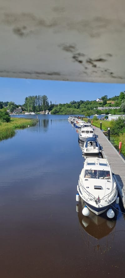 The image shows a view of a calm river or canal with several boats docked along a wooden pier. The boats are of various sizes and designs, mostly white with blue accents. The closest boat has the name "La Vida Loca I" written on its side. The pier extends into the distance, lined with more boats. On the left bank of the river, there is lush green grass and vegetation, with some houses and trees visible in the background. The sky is clear and blue, suggesting a sunny day. The top portion of the image is obscured by what appears to be a textured, light-colored surface with dark, cloud-like markings, possibly the underside of a roof or canopy. The scene is likely a marina or a section of a waterway where boats are moored. The location is identified as being in Enniskillen, United Kingdom.