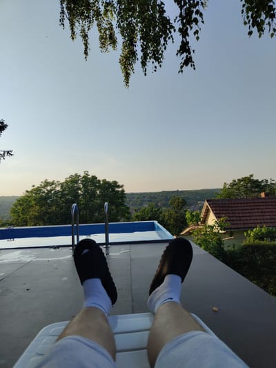 The image captures a first-person perspective from a relaxed, seated position, overlooking an outdoor scene with a pool. In the immediate foreground, the lower legs of an individual are visible, clad in light grey shorts or sweatpants, white socks featuring a dark emblem near the ankle, and dark, slip-on sandals. The individual is occupying a white plastic lounge chair, a portion of which is visible at the bottom center of the frame.

Directly ahead and slightly to the left, a rectangular, above-ground swimming pool is present, featuring a light blue interior and a lighter blue border. Two chrome-finished handrails are positioned at the edge of the pool, extending into the water. The flat surface surrounding the pool and supporting the lounge chair is dark grey or black, appearing to be a composite deck or roof material, with visible water droplets near the pool's edge.

Beyond the pool, a landscape of dense green trees forms the midground and extends into the background, suggesting a heavily vegetated or forested area. To the right of the pool, a section of a residential structure with a reddish-brown tiled roof is partially visible, nestled among the trees. The distant background reveals a sprawling panorama of a settlement or village situated on undulating hills, composed of multiple buildings under a clear, pale blue sky. Overhanging green foliage from a tree drapes into the top portion of the frame. The scene is illuminated by natural light, consistent with daytime. The image provides no specific visual markers to confirm the location as Pisanets, Bulgaria.