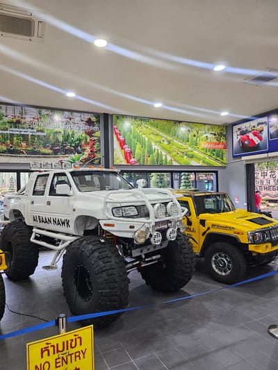 The image shows the interior of what appears to be an exhibit space, likely at Nong Nooch Tropical Garden in Na Chom Thian, Thailand. Dominating the foreground are two large, modified vehicles: a white monster truck on the left with extremely oversized tires and a yellow Hummer on the right, also with large tires. The white truck has "ALBAAN NAWK" written on its side and a license plate with Thai characters and the number "8888". The yellow Hummer has "BFGoodrich" branding and "ALL HUMMER" on its hood.
Behind the vehicles, a large mural depicts a lush, green Italian garden landscape with pathways and water features, labeled "Italian Garden". To the left of this mural is another mural of a "Bromeliads Display Garden" with hanging plants. Above the vehicles and murals, the ceiling features recessed lighting and an air conditioning unit.
A blue tape barrier is placed on the floor in front of the vehicles, and a yellow "NO ENTRY" sign with Thai text is visible in the lower left corner. The flooring appears to be dark grey tiled. In the background, through windows or open doorways, glimpses of more greenery and trees can be seen, suggesting an outdoor area connected to the exhibit. A person in a red shirt is partially visible in the background on the right, near a doorway. The overall scene suggests a display intended to showcase these vehicles within a themed exhibition.