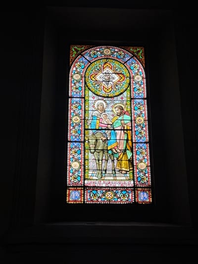 The image shows a stained glass window in a church located in the city of Collbató, Spain. The window depicts a religious scene with three figures: a woman holding a baby, a man standing beside them, and a donkey. The woman and baby are positioned on the donkey, and the man is walking alongside. The figures are dressed in traditional religious attire, with flowing robes in various colors. The stained glass is intricate, featuring detailed patterns and vibrant colors, including blues, reds, yellows, and greens. The scene appears to be a biblical narrative, possibly the Flight into Egypt. The window is arched at the top and framed by a decorative border with geometric and floral motifs. The background of the window is dark, suggesting it is lit from within or by external light sources. The surrounding environment appears to be a dimly lit interior space, consistent with a church.