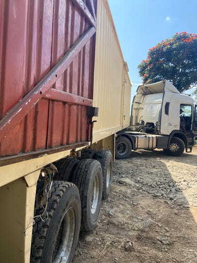 The image depicts two semi-trucks parked on a dirt road in an outdoor environment. The truck on the left has a red cargo container with visible metal bracing. The truck on the right is a white cab-over tractor unit, its driver's side door is open, and a person is visible inside the cab. Behind the trucks, a tree with vibrant orange flowers is in bloom, and the sky is clear blue with a few scattered clouds. The ground is covered in gravel and dirt. The scene appears to be in a rural or industrial area, possibly a transportation hub or construction site. The lighting suggests it is daytime.