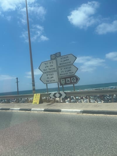 This is a medium shot of a road with directional signs and the ocean in the background. The signs are white with black text in Hebrew and English, directing to Mikhmoret, Mevo'ot Yam, Bet Yanay, and Ne'urim. Some signs also have pictograms indicating beach activities. There is also a yellow sign with Hebrew text listing prices for food items like "Gechon" and "Kubana". The scene appears to be in Israel, given the Hebrew text. The weather is clear with a bright blue sky and scattered white clouds. The ocean is visible behind the signs, and there is a parking lot with many cars along the coast. The overall setting is a roadside with a view of the sea, likely a popular coastal area. No people are prominently visible, but the presence of a parking lot suggests activity in the area. The time of day appears to be daytime due to the bright sunlight.
