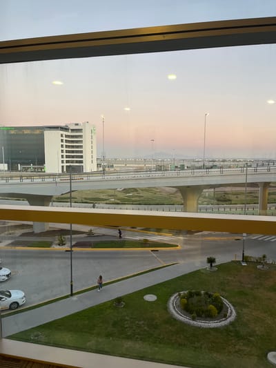 The image depicts an exterior view, likely from an elevated window, overlooking a modern airport environment at twilight, specifically the Felipe Ángeles International Airport (AIFA) near San Miguel Jaltocan, Mexico. The sky transitions from a light blue at the top to pale pink and orange closer to the horizon.
In the immediate foreground, a metallic horizontal window frame (gold and dark gray) is visible, with interior light reflections on the glass.
Below the window frame, the ground level features a structured landscape of concrete walkways, paved roadways with yellow curb markings, and green grass patches. Two white sedans are parked on a paved area to the lower left. A female individual in a light-colored top and jeans walks on a concrete sidewalk towards the lower right. Further into the midground, another person wearing a dark top, red pants, and a dark backpack walks on a sidewalk adjacent to a landscaped area. Landscaping includes small circular garden beds bordered by white pebbles, containing various low-lying plants and small palm trees, along with agave-like succulents. Several illuminated streetlights are interspersed throughout the scene.
Dominating the midground is a multi-lane concrete overpass or elevated road, supported by substantial concrete pillars. It features dark railings along its length.
In the background, beyond the overpass, two prominent buildings are visible. To the left, a large, modern structure with a dark glass facade and a green "Holiday Inn Express & Suites" logo is present. Adjacent to it on the right is a white, multi-story building with numerous windows and balconies, also featuring a green "Holiday Inn" logo. Further into the distance, additional elevated structures and a flat expanse, indicative of airport infrastructure like runways or taxiways, are visible, dotted with numerous distant lights. Faint mountains or hills are silhouetted against the horizon.
