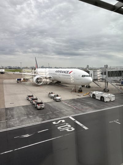 The scene depicts an Air France Boeing 777-300ER aircraft, with partial registration "NR" visible near its nose, parked at a gate on the tarmac of Tokyo Haneda Airport (HND) in Ota, Japan. A passenger jet bridge is connected to the aircraft's forward left door. Ground support equipment includes a large white "tvd" pushback tug positioned near the front landing gear and a yellow-orange service vehicle partially visible by the left engine. On the tarmac, a train of four white baggage carts towed by a small white tractor is parked, alongside two additional baggage carts with their respective tractor. The foreground tarmac features white double lines and a prominent white "STOP" marking. A distant city skyline is visible in the background under an overcast sky. No individuals are discernible, and no active movements or interactions are depicted.