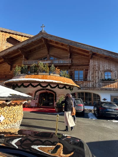 This image depicts the Stanglwirt hotel in Going am Wilden Kaiser, Austria, on a clear, sunny day. The hotel is a large, multi-story structure constructed primarily of natural wood in a traditional Alpine style, featuring a steep gabled roof topped with a decorative cross and multiple wooden balconies. A prominent semi-circular entranceway with a conical wooden shingle roof is visible in the center, adorned with green garlands. A red carpet extends from the entrance onto the paved forecourt. To the immediate left, a smaller wooden structure with a snow-covered roof holds stacked firewood.
In the mid-ground, a woman with dark hair, dressed in a dark vest over a light top, a long cream-colored skirt, and dark heeled boots, walks with her back to the camera, carrying a light brown shoulder bag. A second person in dark clothing is partially obscured behind her, to her right. Two dark-colored vehicles are parked on the right side of the paved area; one is a black Audi SUV with license plate "ERS 3569". The ground shows patches of snow and ice, contrasting with the dry asphalt. Long shadows are cast across the pavement. The bottom foreground reflects the surface of a black vehicle. The sky is bright, clear blue.