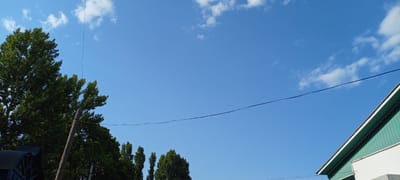 A clear blue sky with scattered white clouds is visible above a section of a green-sided building with a white trim, seen from a low angle. A thin, dark wire runs diagonally across the sky. On the left side of the image, the upper branches of several large green trees are visible, with one prominent tree trunk made of rough, weathered wood extending upwards, seemingly a utility pole. There are also some smaller trees further back. The setting appears to be outdoors, possibly in a residential or rural area within the city of Kirsanov, Russia, judging by the presence of utility poles and trees. The overall scene is bright and suggests daytime, with good visibility.