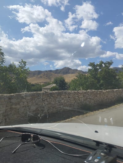 The image shows a view from inside a vehicle, looking out towards a rural landscape in O'xum QFY, Uzbekistan. A stone wall runs horizontally across the mid-ground, with trees and vegetation behind it. A house with a sloped roof is visible nestled among the trees. In the background, rolling hills and mountains are seen under a blue sky with scattered white clouds. The foreground shows a portion of the vehicle's dashboard, with a pair of sunglasses resting on it. The overall scene depicts a sunny day in a rural setting.