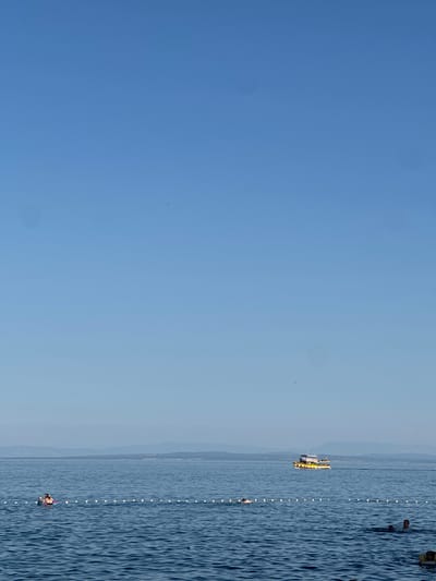 The image depicts a serene view of the Adriatic Sea near Lovran, Croatia. The sky is a clear, bright blue with a few distant, faint smudges. The sea is a deep blue with gentle ripples. A white buoy line stretches across the water, marking a swimming area.
In the distance, a yellow and white tourist boat with multiple decks is cruising across the sea. On the left side of the frame, a person is relaxing on an inflatable tube within the marked swimming area. Further into the water, several other individuals are visible swimming, with some partially submerged. The distant horizon shows a faint outline of a coastline with hills or mountains, suggesting the presence of land beyond the sea. The overall atmosphere is calm and recreational, typical of a summer day at the seaside.