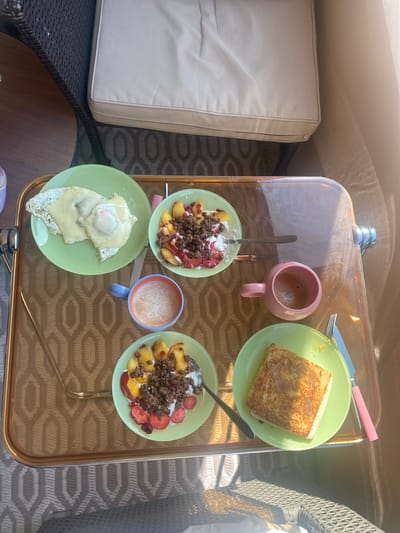 The image shows a top-down view of a breakfast spread arranged on a glass-topped table. The table is set on what appears to be a patio or balcony, with patio furniture including a beige cushion seat and woven chairs visible. The food items include:

1.  A plate with two fried eggs, possibly topped with melted cheese.
2.  Two bowls containing fruit, granola, and possibly yogurt. The fruit includes peaches and strawberries, with what appears to be pomegranate seeds.
3.  A slice of toasted bread, likely French toast, served on a green plate with a knife and fork.
4.  Two mugs with a beverage, one pink and one blue.

The color palette is predominantly pastel, with the green plates and pink and blue mugs contrasting with the brown tones of the table and the furniture. The scene appears to be set in daylight, with natural light casting shadows and highlighting the food. The location in Monthey, Switzerland is not evident from the image.