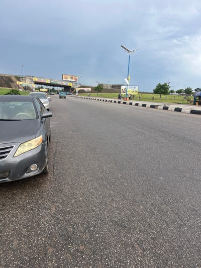 The image depicts a street scene in Osogbo, Nigeria, under a cloudy sky. The foreground is dominated by a gray sedan parked on the side of the road. The road is paved with asphalt and marked with black and white stripes along the shoulder. In the background, there is a green field with scattered trees and a few people. Several billboards are visible, displaying various advertisements. A unique lamppost with three blades on top is also present.