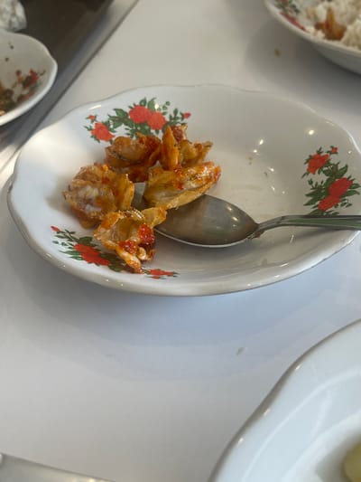 A close-up shot focuses on a white ceramic bowl adorned with a floral pattern of red flowers and green leaves. Inside the bowl, a serving of what appears to be fried fish or shrimp is visible, coated in a reddish sauce. A metal spoon rests within the bowl, partially submerged in the food. The bowl is placed on a white surface, likely a table, with other white dishes or surfaces visible in the background, suggesting a dining setting. The lighting is bright, indicating an indoor environment, possibly a restaurant or home. The scene is static, with no activity depicted beyond the presentation of the food. The overall impression is of a simple, appetizing dish. The location context of Medang, Indonesia, might suggest a local culinary specialty.