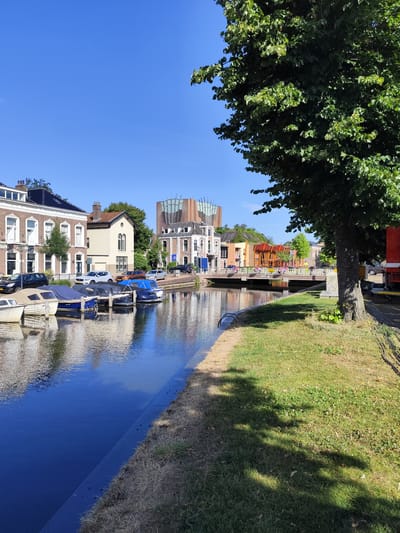 This media file captures a sunny day scene in Sneek, Netherlands, featuring a canal lined with boats and buildings. The canal reflects the blue sky and the facades of the charming European-style houses and modern architecture. Several boats are moored along the canal, some covered with tarpaulins, indicating a peaceful setting.

In the background, a distinctive modern building with a glass facade stands out amongst the traditional architecture. A bridge spans the canal, with people visible on it, suggesting a lively area. The scene is bathed in bright sunlight, casting shadows from the trees and buildings, indicating it is likely midday. The weather appears clear and warm, with a vibrant blue sky.

The foreground is dominated by the calm, reflective surface of the canal and a grassy embankment. A large tree with lush green foliage partially obscures the view on the right side of the image, its shadow stretching across the grass. The overall atmosphere is serene and picturesque, showcasing a blend of historic and contemporary elements in a waterside urban environment. No visible text is present in the image.