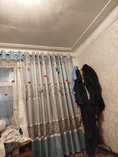 In a domestic interior in Osh City, Kyrgyzstan, the scene is dominated by a set of patterned curtains, featuring a blue and gold motif, drawn across a window. Several small, artificial butterflies in various colors, including red, blue, and orange, are affixed to the curtains and a sheer lace overlaying the window. To the right of the curtains, a collection of clothing, including dark jackets and lighter garments, is draped over a metal rack. In the lower left corner, a pile of laundry rests on a wooden surface. The room's walls are textured, and the ceiling shows signs of age, with some discoloration and imperfections. The overall impression is of a lived-in space, likely a bedroom or living area.