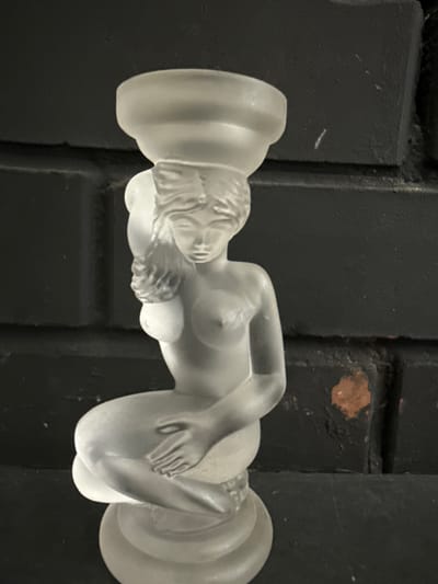 The image shows a frosted glass figurine of a nude woman sitting with her legs crossed and her hands resting on her knees. She has her head tilted down and appears to be gazing at her hands. A bowl-like structure rests on her head. The figurine is positioned in front of a dark, textured brick wall. The lighting is soft, casting subtle shadows and highlighting the frosted texture of the glass. The overall setting appears to be indoors, possibly on a shelf or mantelpiece. There is no indication of the specific location within the city of Хохряки, Russia.