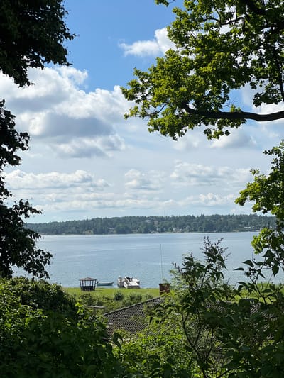 The image depicts a serene waterside scene in Österskär, Sweden, framed by lush green foliage from trees in the foreground. The expansive, calm body of water dominates the mid-ground, with a distant tree-lined shore visible. On the water's edge, a wooden dock with lounge chairs extends outwards, and a small, light-colored boat is moored nearby. A hexagonal gazebo with a dark roof stands on the grassy embankment, further enhancing the tranquil atmosphere. The sky above is a vibrant blue, dotted with fluffy white clouds. The overall impression is one of peaceful natural beauty and summer relaxation.