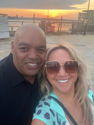 A selfie image captures a bald-headed man and a blonde woman, both smiling broadly. The woman is wearing sunglasses and a light blue top with dark blue spots. Behind them is a view of the Mersey River at sunset from a location in Liverpool, UK. A metal fence and industrial structure with a "DANGEROUS STRUCTURE KEEP OUT" sign are also visible in the background.