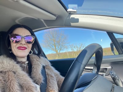 A woman with dark hair, wearing sunglasses, a pearl necklace, and a fur coat, is positioned in the driver's seat of a car. The scene is viewed from within the vehicle, with the steering wheel visible. A tree, reflecting in the windshield, indicates an outdoor environment. Given the city name Roodt-sur-Syre, Luxembourg, it is likely the photo was taken there, though there is no specific identifying information visible to confirm the exact location.