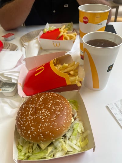 An overhead shot captures a close-up of a McDonald's meal laid out on a white table in Pontecagnano Faiano, Italy. In the foreground, a hamburger with a sesame seed bun, overflowing with shredded lettuce and a slice of cheese, is presented in a white cardboard box. To the right of the hamburger, a white cup with a yellow stripe contains a dark beverage. Behind it, a larger Coca-Cola cup also features the distinctive yellow stripe and the Coca-Cola logo. Further back, two red and white McDonald's french fry containers are visible, one partially filled with golden fries. A small red packet, likely containing sauce, sits to the left of the fries. White napkins are scattered around the food items. In the background, the arms and torso of a person wearing a dark shirt are visible, suggesting they are seated at the table. A receipt, partially visible on the right side of the image, indicates a transaction took place.