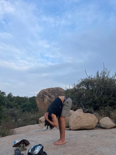 The image captures an outdoor scene on a rocky terrain in Sanapur, India, under a partly cloudy blue sky. A male individual, barefoot, stands in the center-right, bending forward at the waist with his head down and hands clasped behind his neck or head, appearing to stretch. He is wearing a black t-shirt and plaid shorts, with a "Wilson" brand waistband visible. To the lower left of the individual, on the flat stone ground, are several items: a partially filled clear plastic water bottle, a crumpled brown fabric item, a pair of dark sandals, and a black and white motorcycle helmet. The background features large, rounded brown boulders, dense green shrubs, and trees, indicating a natural, unurbanized landscape.