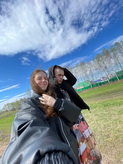 Two people are lying on a blanket outdoors. A woman with long brown hair and a black leather jacket is in the foreground. Behind her is a man in a black hooded jacket and a plaid shirt. They appear to be on a grassy area. Beyond the grassy area are trees. The sky is blue with white clouds. The location is Ulukulevo, Russia.