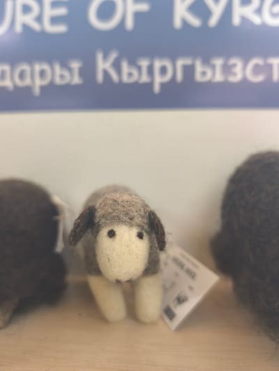 The image displays a close-up, shallow depth-of-field shot of a small, handcrafted felted sheep toy. The sheep, positioned centrally and in sharp focus, is predominantly off-white with light brown detailing on its head and ears, and small dark bead eyes. It rests on a light wooden surface.

On either side of the central sheep, out of focus, are parts of two other dark brown, fluffy textile items, likely similar felted or knitted creations. A small, white product tag is attached to the central sheep, and although blurry, the word "WOOL" can be faintly discerned on it, indicating the material.

In the blurred background, a blue sign with white text is visible. The top line of English text appears to read "URE OF KYRGI," strongly suggesting "CULTURE OF KYRGYZSTAN." Below this, a line of blurred Cyrillic text can be partially deciphered as "дары Кыргызст," likely translating to "gifts/medicines of Kyrgyzstan" or part of a larger phrase referencing Kyrgyzstan.

The scene appears to be an indoor display of local handicrafts, specifically wool products, from the Кашат region of Kyrgyzstan. The lighting is soft, and the overall impression is one of artisan work and cultural representation.