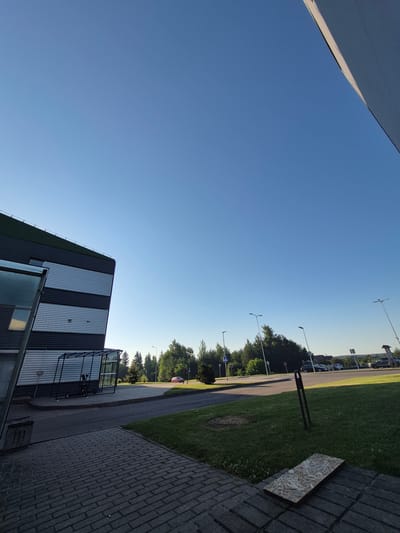 The image depicts an outdoor scene in Pabradė, Lithuania, with a modern building on the left side. The building has a dark, corrugated facade with horizontal light gray stripes. Adjacent to the building is a covered shelter with a metal frame and glass panels, housing two electric scooters. A paved walkway made of gray bricks leads away from the building and into the foreground. To the right of the walkway, a patch of green grass is visible, with a piece of oriented strand board lying on the bricks. Beyond the building and walkway, a road curves into the distance, lined with trees and streetlights. Several cars are parked in the background. The sky is clear and blue, suggesting it is daytime. The scene appears to be a commercial or industrial area, possibly a business park or a community center, due to the modern architecture and the presence of parking facilities. There are no people visible in the image.