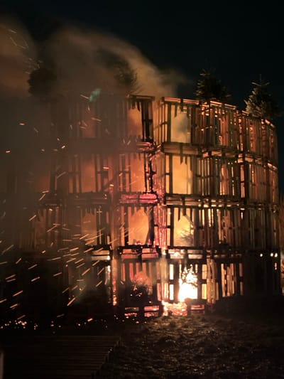 A towering structure built from wooden pallets is ablaze at night. Flames lick through the grid-like construction, while sparks shoot out from the burning wood. Smoke billows from the structure, obscuring parts of it. Small fir trees are perched at the top of the construction. The scene is dimly lit, with the fire providing the primary source of illumination.