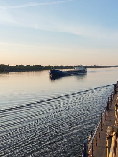 In the city of Bol'shoi Dvor, Russia, a wide, calm river reflects the soft light of a sunset sky. A long, blue cargo ship is moving down the river, leaving a visible wake. On the right side of the image, a portion of another vessel is visible, showing its deck, railings, and some equipment. The riverbanks are lined with lush green trees. The sky is a gradient of pale blue and orange, with a few wispy clouds.