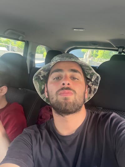 A man wearing a camouflage bucket hat and a dark t-shirt is in the front passenger seat of a car. He is looking at the camera with his lips pursed. A person in a red shirt is visible in the back seat to the left. The car's interior, including seats and ceiling, is visible. Outside the car windows, trees and buildings can be seen. The image appears to have been taken during the daytime. The location is Stepantsminda, Georgia.