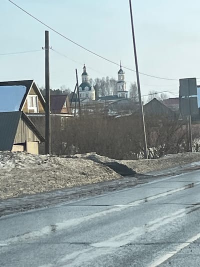 The scene depicts a street view in Чутырь, Russia, under clear skies, indicative of late winter or early spring. In the foreground, a two-lane asphalt road with faint white markings is partially covered by ice and melted snow. Adjacent to the road, banks of dirty snow and exposed soil are present. The midground features a cluster of residential structures, including a two-story wooden house with a snow-covered gable roof on the left, and various smaller buildings. Numerous leafless trees and shrubs occupy the central midground. Multiple utility poles and overhead power lines intersect the frame. Dominating the background are two distinct Orthodox church structures visible above the tree line: a church with a green dome and white facade, and a taller, lighter-colored bell tower with a spire. No human activity or individuals are observed. Lighting suggests bright daytime conditions.