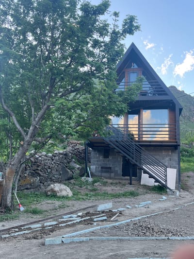 In Sioni, Georgia, a modern A-frame cabin under construction stands amidst a rustic environment. The cabin features a dark metal roof and exterior trim, with large windows reflecting the cloudy sky. A dark metal staircase provides access to the upper level, which has a balcony. The cabin's foundation appears to be constructed with gray concrete blocks. To the left of the cabin, a large tree with lush green foliage dominates the foreground, its branches extending towards the building. Behind the tree, a low stone wall constructed from various-sized rocks separates the immediate yard from a grassy slope. The ground in front of the cabin is a mix of dirt and gravel, with visible construction materials, including concrete blocks, metal bars, and scattered stones, indicating ongoing work. A red-handled broom with a red brush lies on its side near the base of the tree. In the background, a steep, rocky mountain rises under a bright, partially clouded sky. The overall scene depicts a remote, natural setting with a contemporary dwelling in its early stages of completion.