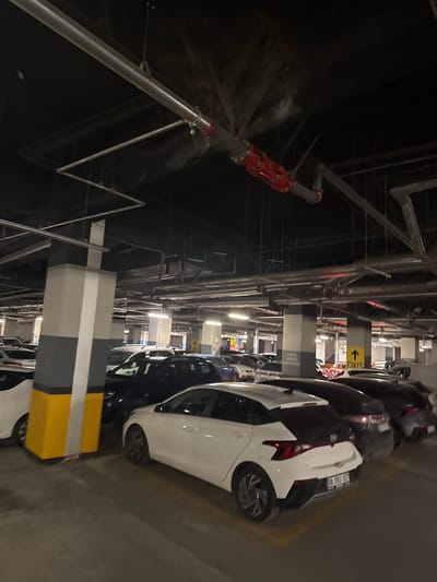 The image shows a multi-story parking garage, likely in Çankaya, Turkey, with cars parked in rows. The lighting is artificial, typical of an indoor parking facility. Overhead, a network of pipes and structural elements are visible, including a red industrial pipe with a cylindrical attachment mounted on the ceiling. Concrete pillars, painted white and gray with yellow base accents, support the structure. The cars are predominantly modern models, in various colors including white, dark blue, and gray. One white car in the foreground is a Hyundai i20, its rear license plate readable as "06 FHJ 637." Signage indicating "GİRİŞ" (Entrance) and "ÇIKIŞ" (Exit) with arrows is visible on some pillars. The overall impression is that of a functional, albeit dimly lit, urban parking area.