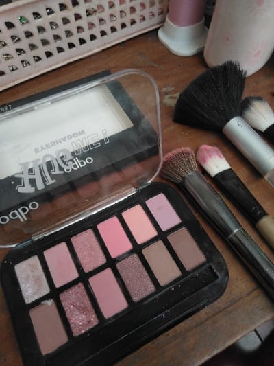 The scene depicts an indoor arrangement of cosmetic items on a wooden surface in Шөптікөл ауылдық округі, Kazakhstan. In the foreground, a black plastic eyeshadow palette is open, revealing ten pans of eyeshadow in various shades of pink, brown, and lavender, with both matte and shimmer finishes. One pan in the lower left corner appears mostly depleted. Directly behind this palette, a second black compact with a clear, partially open lid displays mirrored text "WE! EYESHADOW" and "odbod". To the right of the palettes, five makeup brushes are laid out. These include one large brush with black bristles, one medium brush with light bristles, and three smaller brushes, two of which have pink-tipped bristles. In the upper left background, a segment of a light pink, perforated plastic basket is visible. The upper right background shows two partially visible containers; one is pink with a white base, and the other is a lighter color, possibly white with a floral design. The objects rest on a textured wooden surface. No individuals are present.
