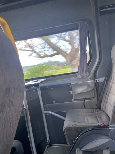 The interior of a bus is seen through a window, revealing a landscape of fields and hills. A partially-drawn curtain is visible on the window. The bus interior includes a seat with a safety restraint system. There is no information to determine the specific city location.
