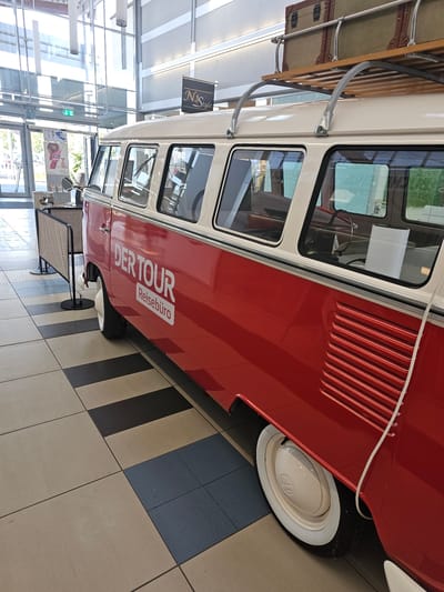 This media file displays a side view of a vintage red and cream Volkswagen van, often referred to as a "Kombi" or "Bus." The van is parked indoors, likely in a public space given the tiled floor with a striped pattern and the large glass windows and ceiling in the background. The lighting suggests it is daytime.

The van is adorned with text on its side that reads "DER TOUR Reisebüro," indicating it is associated with a travel agency. A luggage rack is mounted on the roof of the van, suggesting a theme of travel or a display related to journeys. The van's iconic shape, including the ribbed vents on the side and the white-rimmed wheel, are clearly visible.

Other objects in the scene include what appears to be a woven bench or chair in the foreground and possibly signage or posters on a glass partition further in the background, though these are not clearly discernible. The overall impression is of a well-maintained vehicle presented as an exhibit or a unique promotional tool for the travel agency. The setting is modern and clean, contrasting with the retro design of the van. There are no people visible in the image.