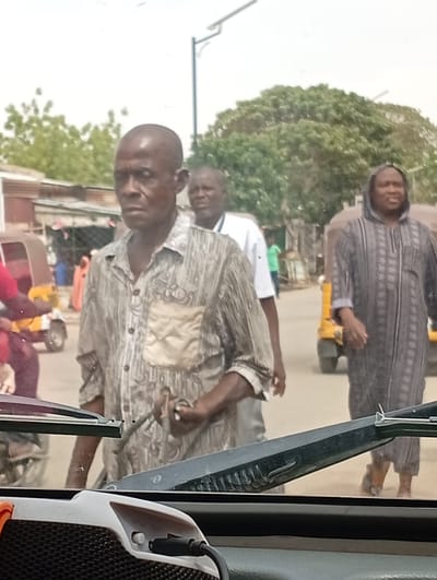 The image captures a street scene in Hotoro North, Nigeria, viewed from inside a vehicle. The foreground features a dark vehicle dashboard with a white and black electronic device, possibly a portable speaker, plugged in via a USB cable and displaying an orange accent. Two dark windshield wiper blades extend horizontally across the lower middle of the frame.
The central subject is a bald, dark-skinned adult male wearing a light-colored, patterned long-sleeved button-up shirt with a lighter rectangular patch on the left chest pocket. He looks slightly to his left (viewer's right) with a neutral expression and holds a dark, indeterminate object in his right hand. Behind him, another dark-skinned adult male in a white short-sleeved shirt is partially visible. To the right of the central figure and further back, a third dark-skinned adult male walks towards the viewer, dressed in a dark, hooded, vertically patterned long garment.
The environment includes an unpaved or lightly paved road. On the left side, a yellow and brown motorized tricycle is partially visible, with a person in a red garment near it. Another yellow motorized tricycle is partially visible on the right side of the road. Buildings with corrugated metal roofing are present on the left side of the street, and dense green foliage from trees dominates the background. A dark street light pole with an extended arm is visible in the upper mid-ground. Additional indistinct figures are present in the background.