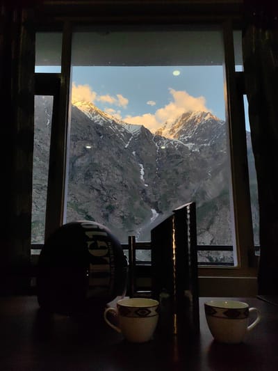 This media file depicts a serene mountain landscape viewed through a window. The scene is set indoors, with the foreground occupied by a dark wooden table. On the table are two white ceramic cups with decorative bands, a dark helmet with "RIDER" and "ROYAL Enfield" visible on it, and a tall, dark, metallic object, possibly a thermal flask.
The window frames a breathtaking view of jagged, rocky mountains partially covered in snow. The peaks are illuminated by the golden light of either sunrise or sunset, casting warm hues on the clouds and snow. Streaks of meltwater or small waterfalls are visible cascading down the mountainsides. The sky is a clear blue with scattered white and orange clouds. The time of day appears to be either early morning or late afternoon, given the lighting on the mountains.
The overall mood is tranquil and contemplative, suggesting a moment of rest and appreciation for the natural beauty of the location. The presence of the helmet and cups hints at a traveler taking a break, perhaps enjoying a beverage while gazing at the stunning vista. The location context provided, Gemur, India, aligns with the mountainous terrain. No specific activity or event is actively taking place, rather it is a static scene of observation and repose.