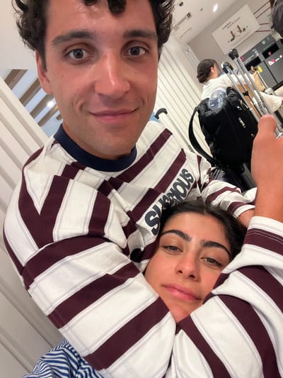 Two individuals, a man and a woman, are captured in a close-up selfie. The man is positioned behind the woman, with his arms around her. Both are wearing striped long-sleeved shirts. The woman's head is resting on the man's shoulder or chest. The background suggests an indoor environment, possibly an airport or a terminal, with luggage and signs visible. The woman's shirt has the word "SUSPICIOUS" printed on it in dark lettering. There are no discernible elements in the image that would place the scene in Mykonos, Greece.