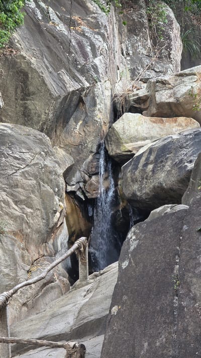 The image depicts a waterfall cascading through a rock formation in Nam Ninh Hoa, Vietnam. The rocks are gray and weathered, with some patches of green moss and small plants growing on them. Water flows through a narrow gap in the rocks, creating a turbulent stream. A rudimentary wooden and rope ladder is visible alongside the water, presumably providing access to the area. No people are visible in the scene. The overall environment appears natural and rugged.
