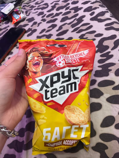 A hand holds a bag of "ХРУСТ team" brand snacks. The bag is primarily red and yellow, with an illustration of a cheering woman with face paint and a soccer ball. The text "ФУТБОЛ ВКУСНЕЕ ВМЕСТЕ" is visible, translating to "Football is tastier together." Below this, "ХРУСТ team" is prominently displayed. The lower portion of the bag is yellow and shows images of two puffed snacks and the text "БАГЕТ" and "«СЫРНОЕ АССОРТИ»," meaning "Baguette" and "Cheese Assortment." The background is a leopard-print fabric. The image appears to be taken indoors, with a mobile phone and other small items visible in the upper left corner. The scene is a close-up of the snack bag held by a person, with no specific indicators of the city of Borisova Griva Massiv, Russia.