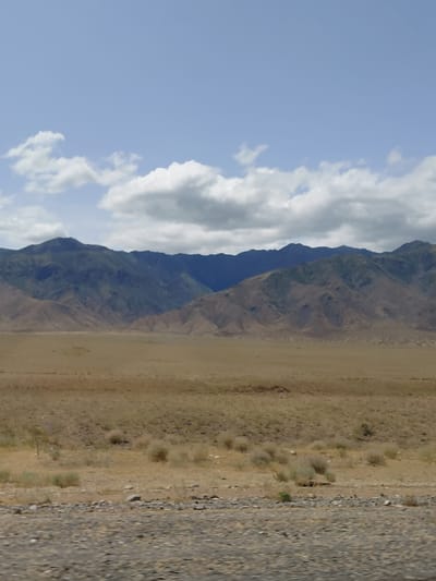 The image displays a wide, panoramic view of a mountainous, arid landscape under a partly cloudy sky. In the foreground, a desolate, rocky plain stretches out, dotted with sparse, dry vegetation. The middle ground is a vast expanse of tan-colored earth, also sparsely covered with low-lying, scrubby bushes. In the background, a range of imposing mountains rises, their slopes displaying a mix of brown and green hues, suggesting varied terrain and vegetation. The sky above is a pale blue, with scattered white and gray clouds that cast shadows on the distant mountains. The overall impression is one of natural, undeveloped wilderness. The location is specified as Ынталы, Kazakhstan.