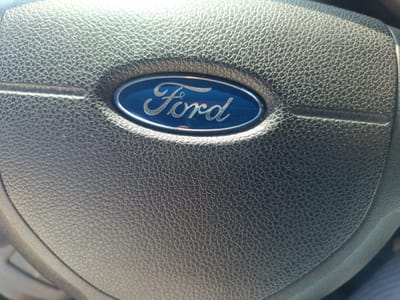 The image shows a close-up of a Ford steering wheel. The Ford logo is prominently displayed in the center, with the word "Ford" written in silver cursive lettering on a blue background, surrounded by a silver oval border. The steering wheel itself is made of black, textured leather. The lighting suggests the photo was taken during the daytime, likely inside a vehicle, with sunlight illuminating the top left portion of the steering wheel. The environment beyond the steering wheel is out of focus, but appears to be the interior of a car. There are no people visible in the image. The image is a static shot and does not depict any action. The location of Chusovoy, Russia, is not discernible from the visual information presented in the photograph.