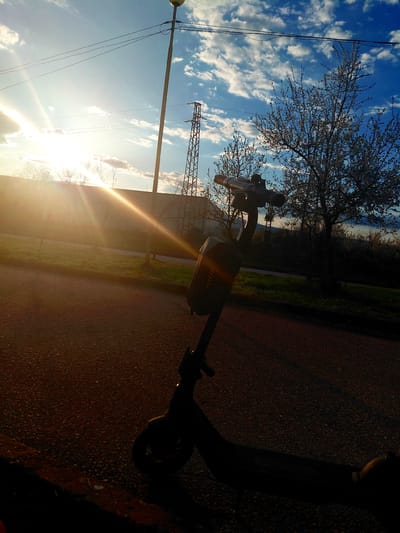 The image, likely taken at ground level, is oriented sideways. The sky is visible in the background, exhibiting a blue hue with scattered white clouds. A tree with bare branches is also present, with power lines strung overhead. A structure, possibly a building, stands in the mid-ground.
In the foreground is the silhouette of a scooter, positioned on what appears to be a paved surface, possibly a road or pathway. The lighting is dramatic, with a bright sunlight shining through, casting strong shadows and creating a lens flare effect. The location is unspecified, and there are no people visible in the frame. The scene is static, without any evident action.
