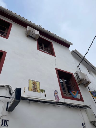 A low-angle exterior view depicts the white-stucco facade of a multi-story building in Buñol, Spain, likely near "El Castillo" as indicated by a prominent street sign. The building features dark red trim around its windows and along the roofline, which is topped with white, possibly terracotta, tiles. A light grey, overcast sky occupies the upper portion of the frame.
Three visible rectangular windows possess dark brown wooden frames; one in the upper left, one in the upper right, and a larger, open window in the mid-right. Two white Kosner brand outdoor air conditioning units are mounted on the facade with wooden brackets; one is positioned above the upper-right window, and another is to the right of the mid-right window. A vertical hairline crack extends down the white wall between the upper and lower sections.
Centered on the lower-mid facade is a rectangular ceramic tile mosaic depicting a crowned and haloed religious figure, likely the Virgin Mary, holding a child, set against a golden background with a yellow border. Below this mosaic, on the left, a dark-colored, rectangular sign bearing the white text "El Castillo" extends from the wall. The house number "10" is affixed below this sign, and the letter "A" is visible further right, below a dense bundle of black utility cables that traverse the facade horizontally. Various decorative elements hang from these cables and window frames: a string of light-colored beads to the left of the tile mosaic, a green crocheted triangular element with tassels, a circular multi-colored (purple, white, blue) crocheted ornament with tassels, and a blue, green, and red crocheted square draped across the mid-right open window. Another yellow and green crocheted item is partially visible on the far right.